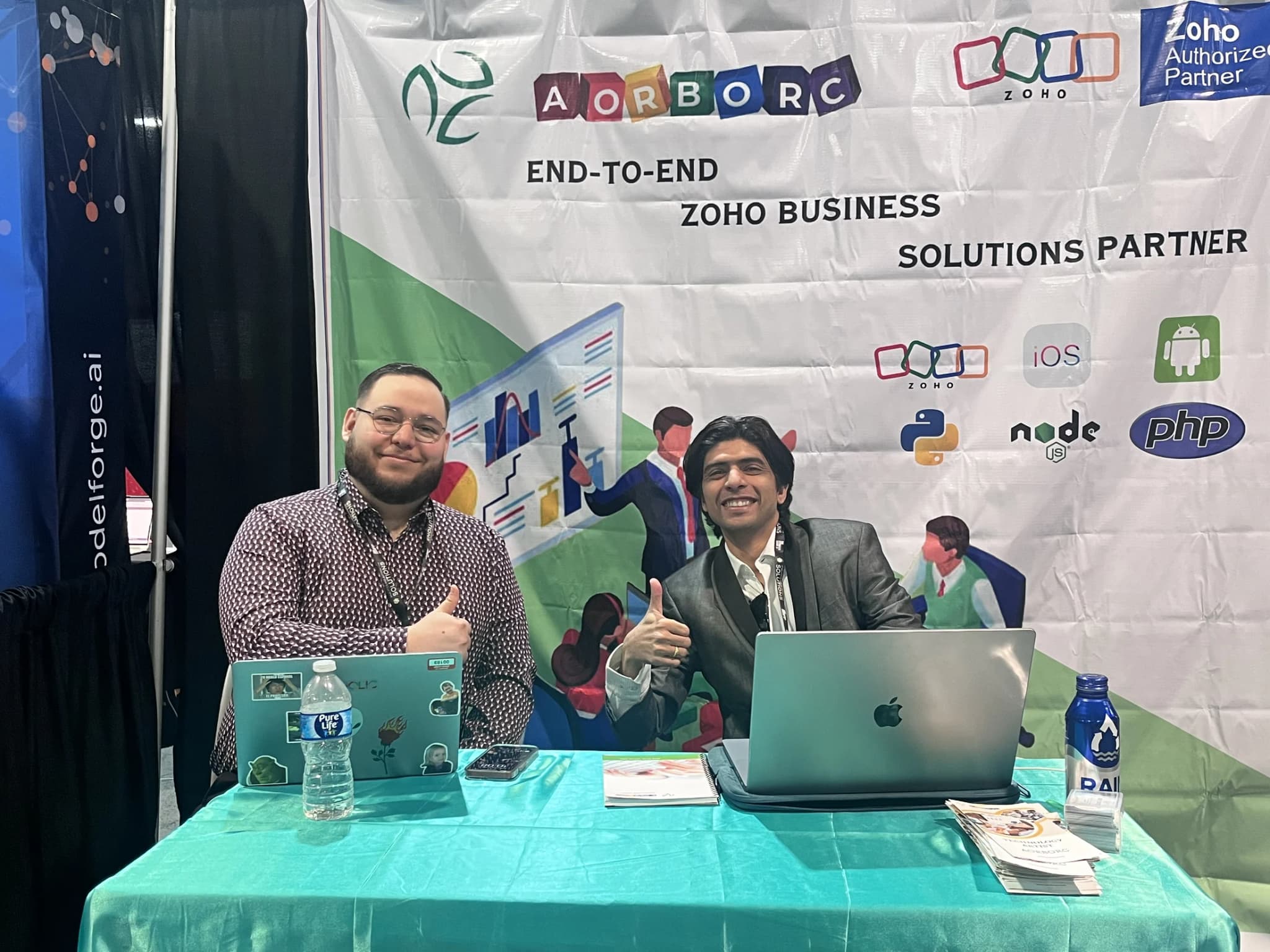 AorBorC Technologies booth at The Business Show, Las Vegas, with Zoho Authorized Partner signage and two team members seated at the demo table.